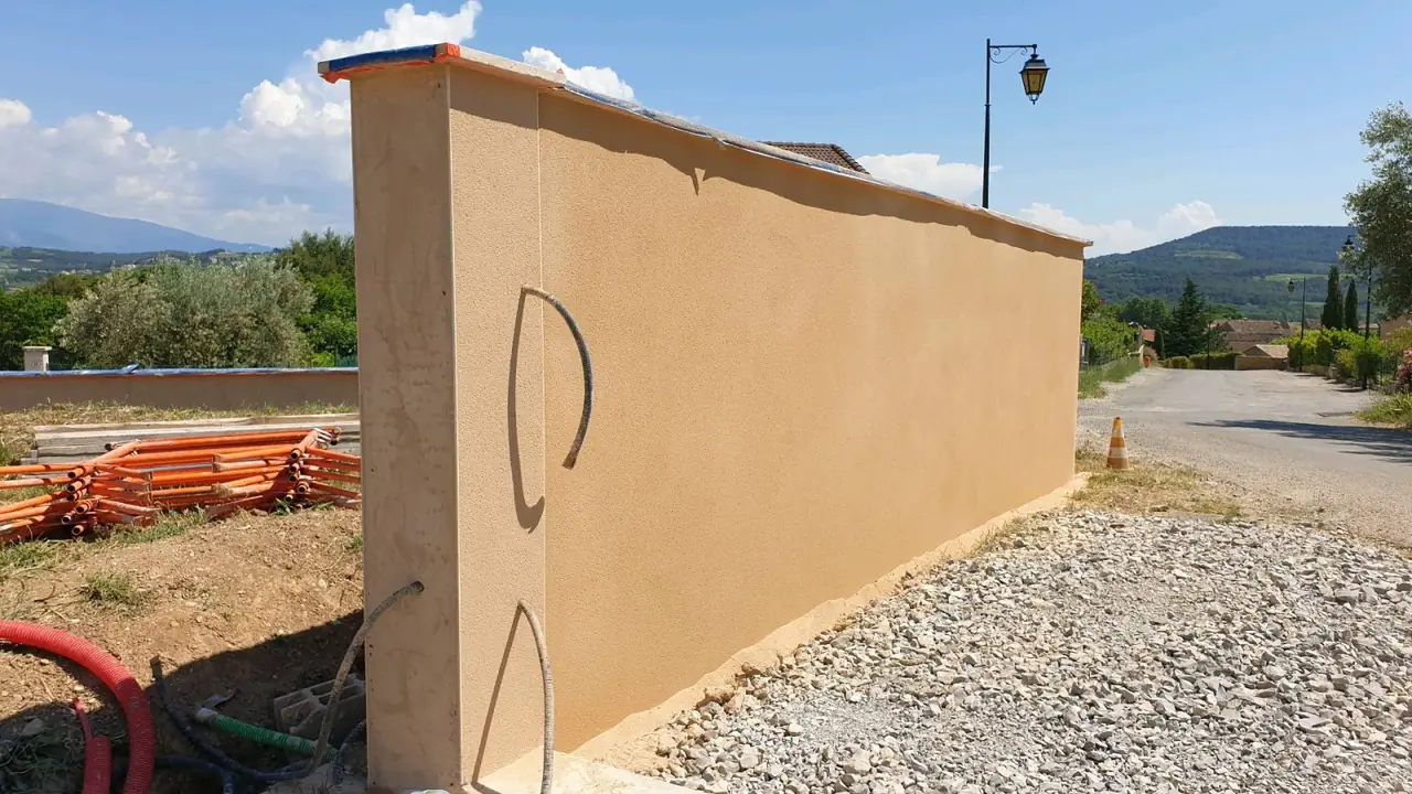 Mur de clôture neuf enduit ocre avec son chaperon, gravier au pied, collines au fond