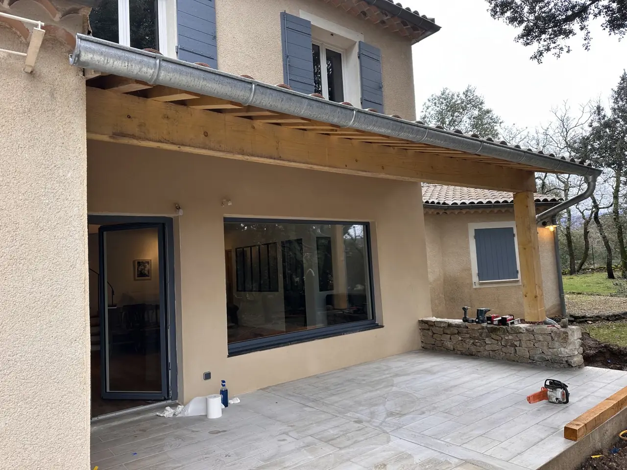 Maison enduite à volets bleus : avancée de toit en tuiles sur poteau bois, terrasse dallée devant une grande baie