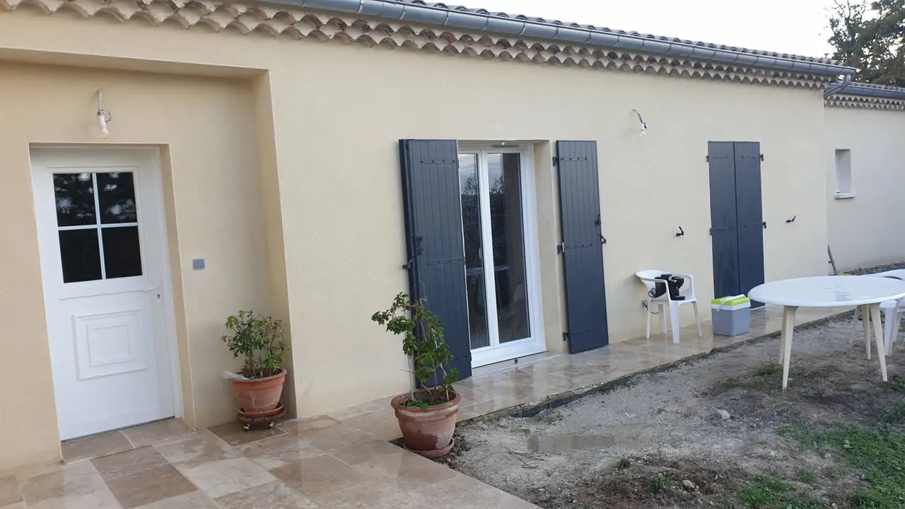 Maison de plain-pied à l’enduit ocre clair, volets bleu-gris, porte d’entrée blanche, terrasse en dalles